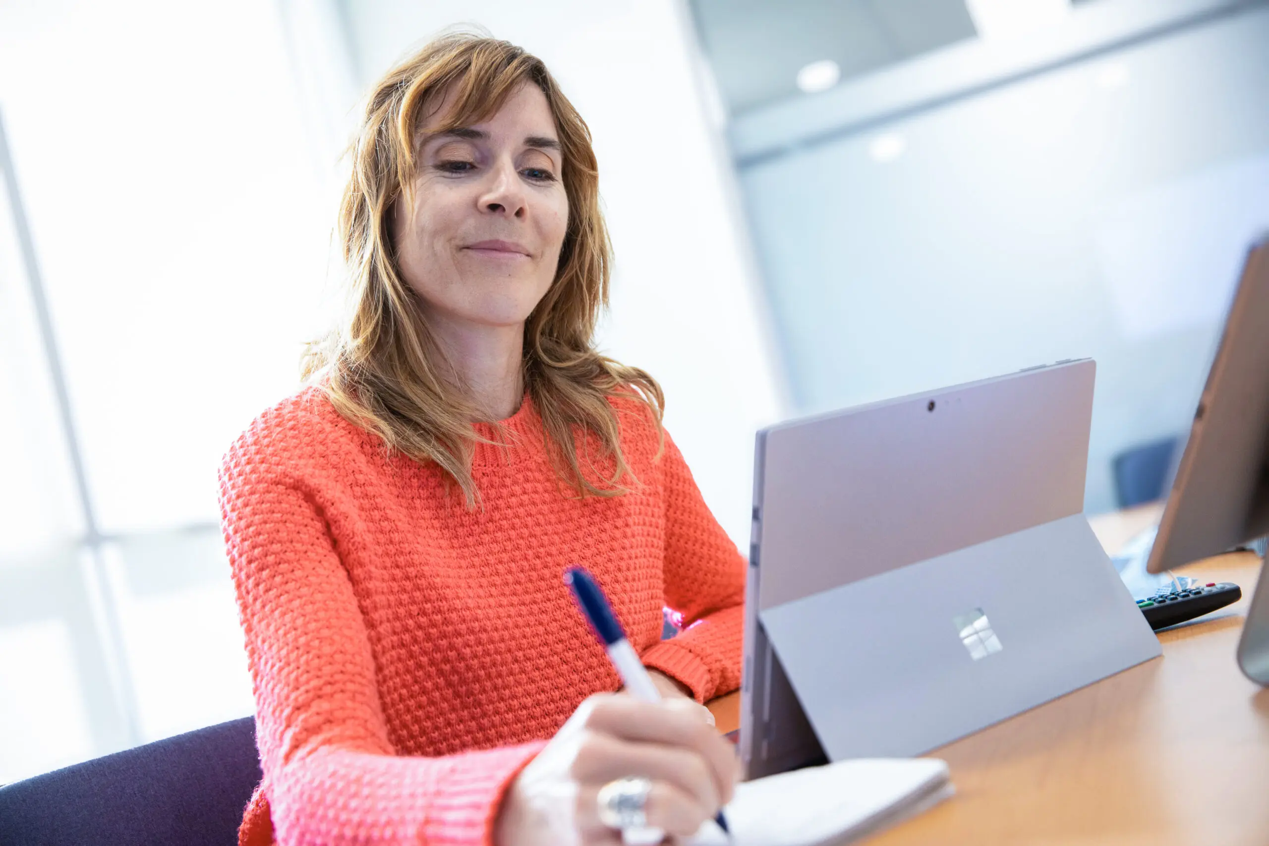 Une femme vêtue d'un pull orange écrit dans un cahier tout en travaillant à un bureau, un ordinateur portable posé devant elle, dans un bureau baigné de lumière.