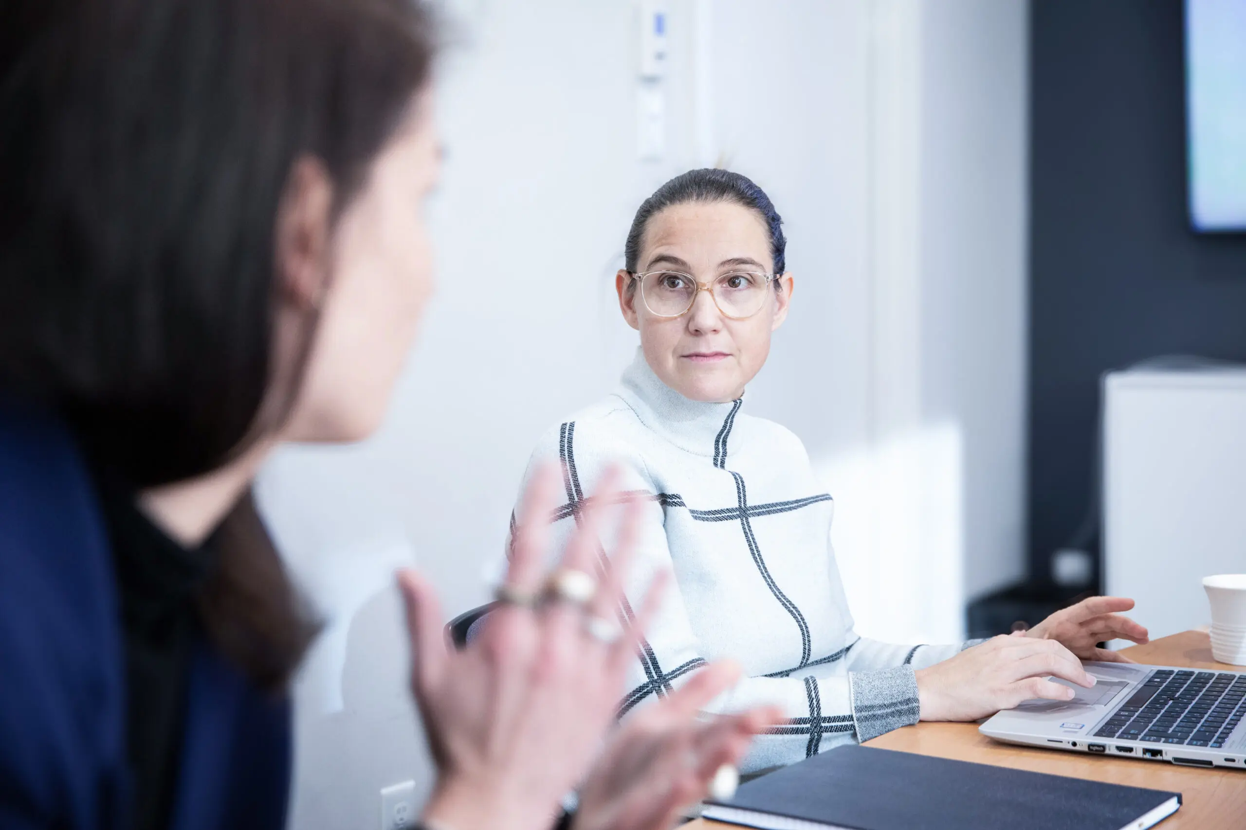 Deux femmes sont assises à une table dans un bureau. L'une d'elles tape sur un ordinateur portable et regarde l'autre, qui parle en accompagnant ses propos de gestes. Un cahier et une tasse se trouvent sur la table. La pièce est lumineuse et moderne.