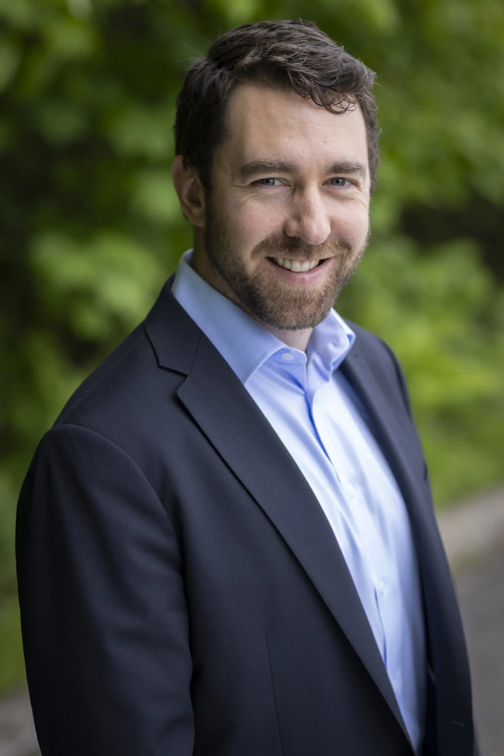 Un homme aux cheveux bruns courts et à la barbe, vêtu d'une veste de costume sombre et d'une chemise bleu clair, souriant en extérieur avec un feuillage vert en arrière-plan.