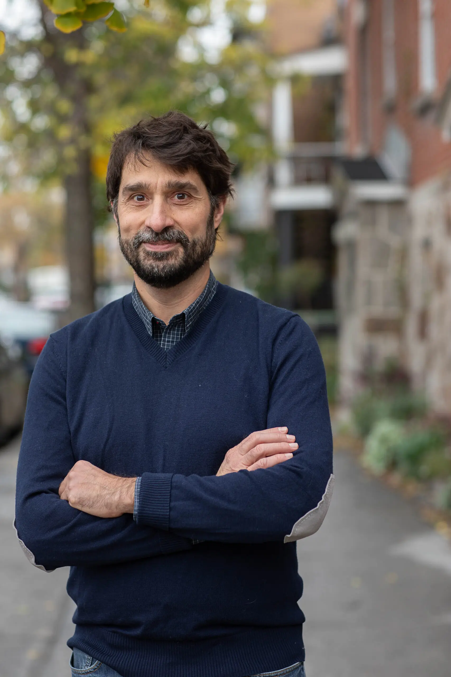 Un homme barbu, portant une moustache, se tient sur un trottoir, les bras croisés, vêtu d'un pull bleu marine. On aperçoit des arbres, des plantes et des immeubles en briques en arrière-plan.