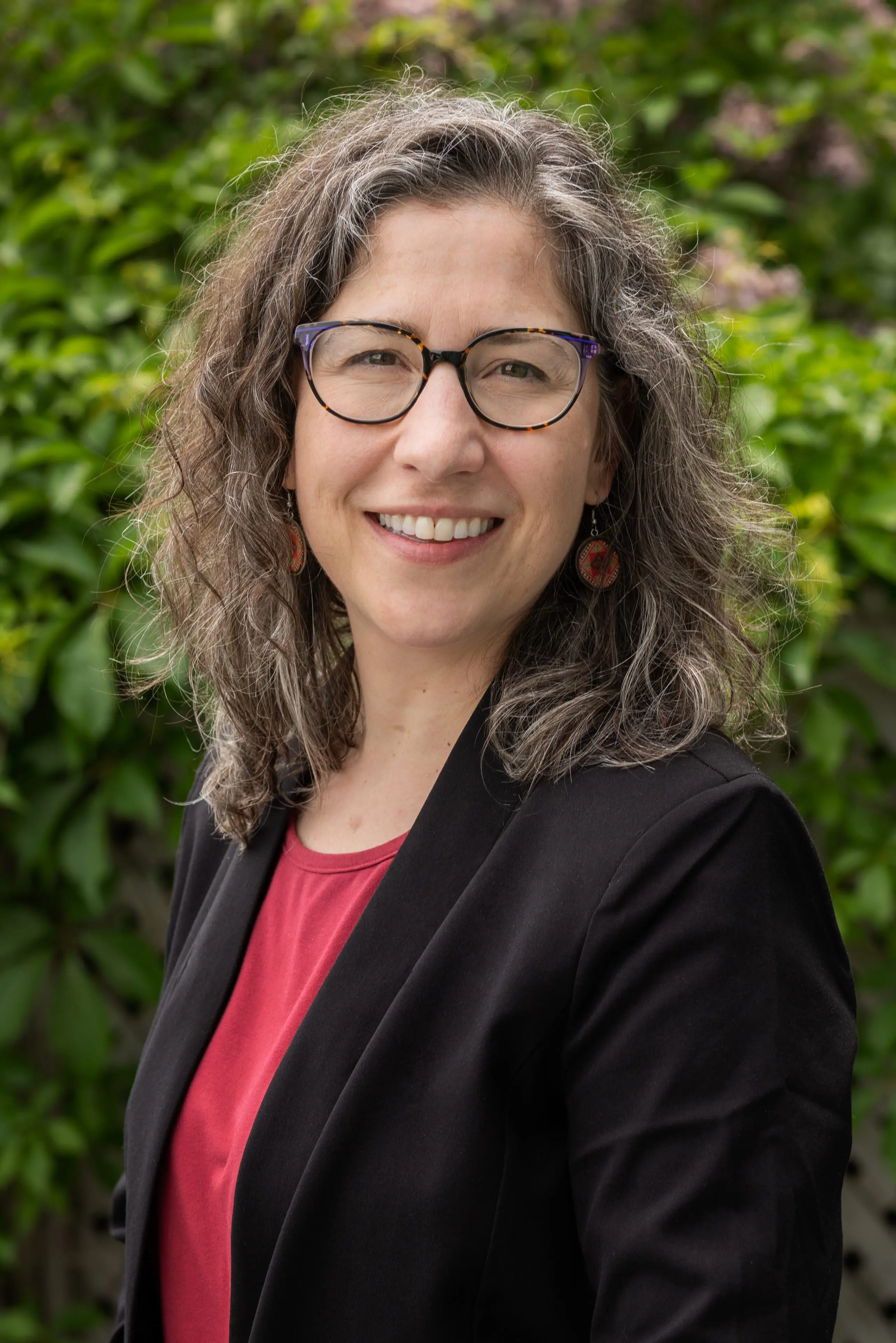 Une femme aux cheveux gris ondulés, portant des lunettes, sourit debout à l'extérieur. Elle porte un blazer noir par-dessus un haut rouge, ainsi que des boucles d'oreilles rondes, tandis qu'un feuillage verdoyant apparaît flou en arrière-plan.