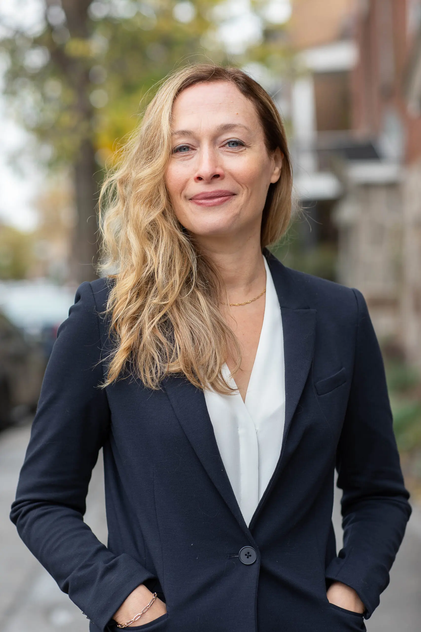 Une femme aux cheveux blonds ondulés, vêtue d'un blazer bleu marine et d'un chemisier blanc, se tient debout sur un trottoir en ville, un doux sourire aux lèvres, les mains dans les poches.
