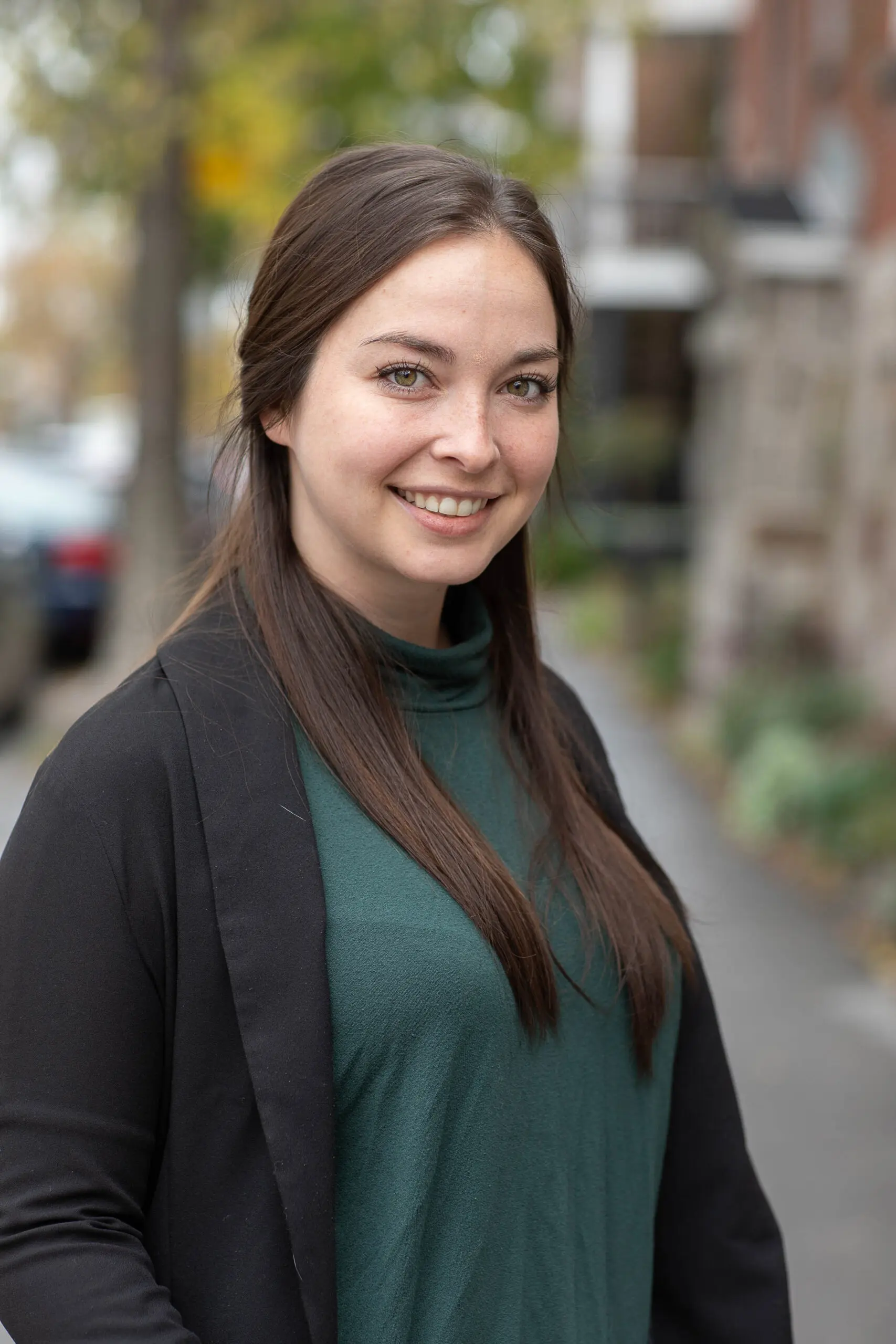 Une femme aux longs cheveux châtains et aux yeux verts sourit à l'extérieur ; elle porte un haut vert foncé et une veste noire. L'arrière-plan est flou et laisse entrevoir une rue bordée d'arbres et de bâtiments.