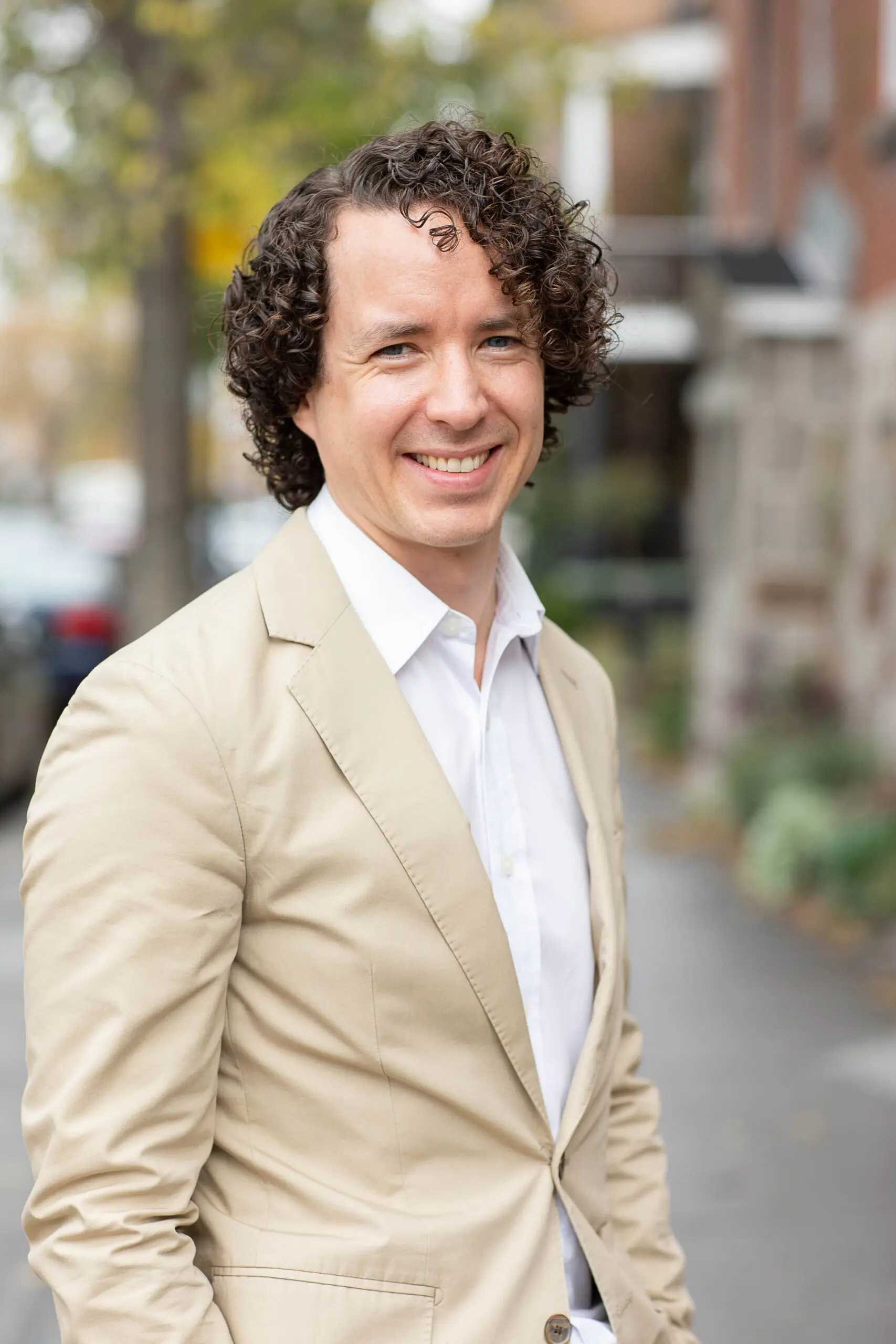 Un homme aux cheveux bouclés, vêtu d'un blazer beige et d'une chemise blanche, sourit, debout sur un trottoir en ville, avec des arbres et des immeubles en arrière-plan.