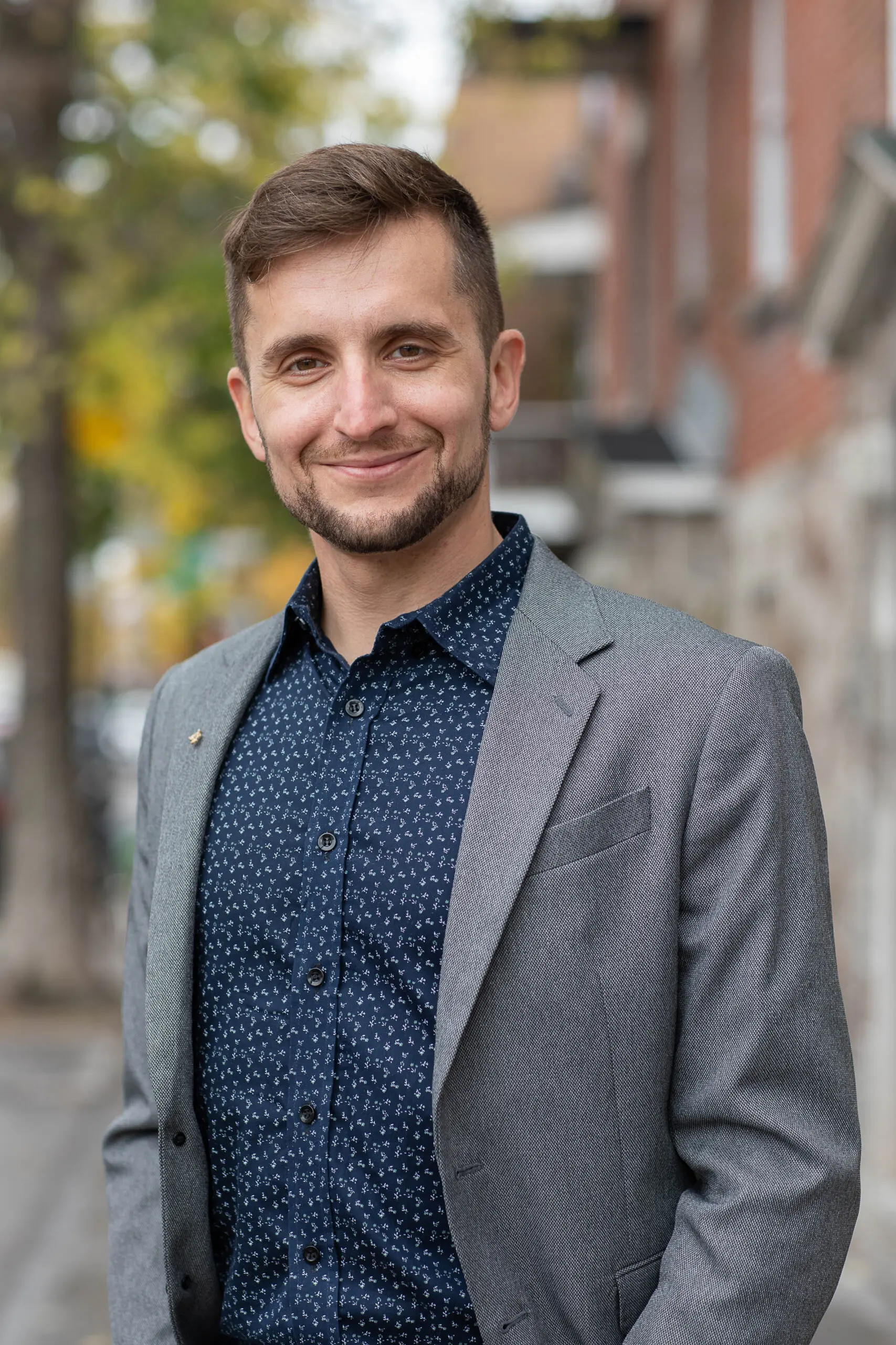 Un homme vêtu d'un blazer gris et d'une chemise bleu marine à motifs sourit à l'extérieur, dans une rue de la ville, avec des arbres et des immeubles flous en arrière-plan.