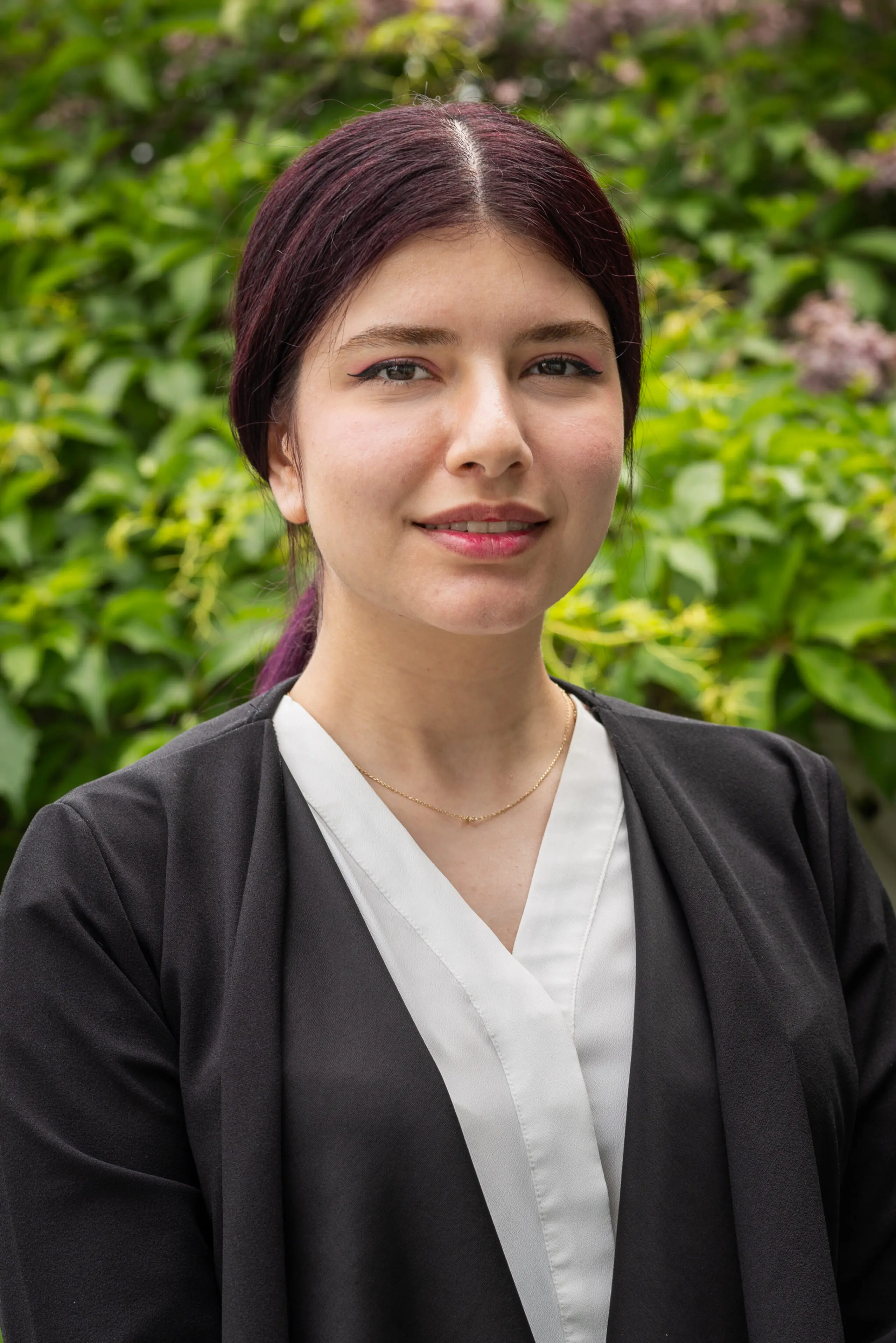 Une femme aux cheveux foncés, vêtue d'un chemisier noir et blanc, esquisse un léger sourire alors qu'elle se tient debout à l'extérieur, devant un feuillage vert.