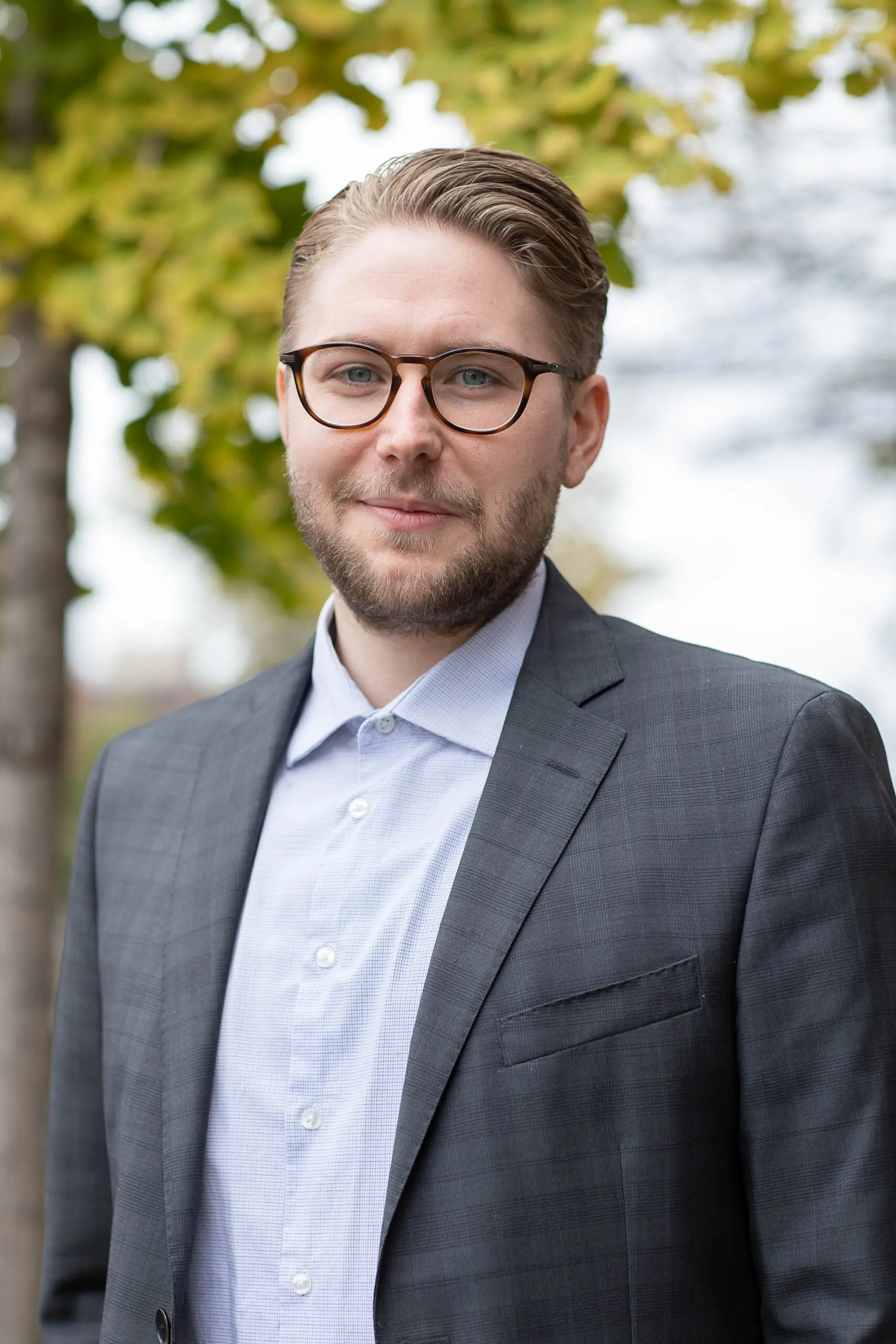 Un homme aux cheveux châtain clair, portant des lunettes et une barbe courte, est vêtu d'un costume gris et d'une chemise claire ; il se tient debout à l'extérieur, devant des arbres aux feuilles vertes et jaunes, et sourit légèrement à la caméra.