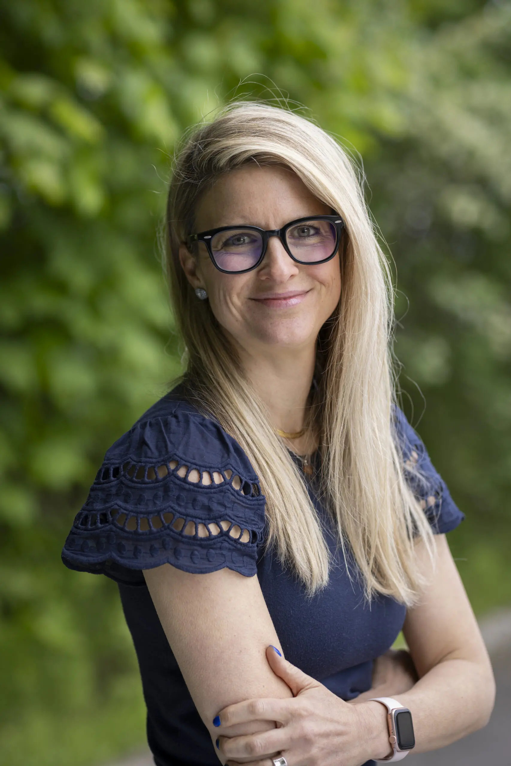 Une femme aux longs cheveux blonds et portant des lunettes se tient dehors, les bras croisés, vêtue d'un haut bleu marine et d'une montre connectée, souriant devant des arbres verts.