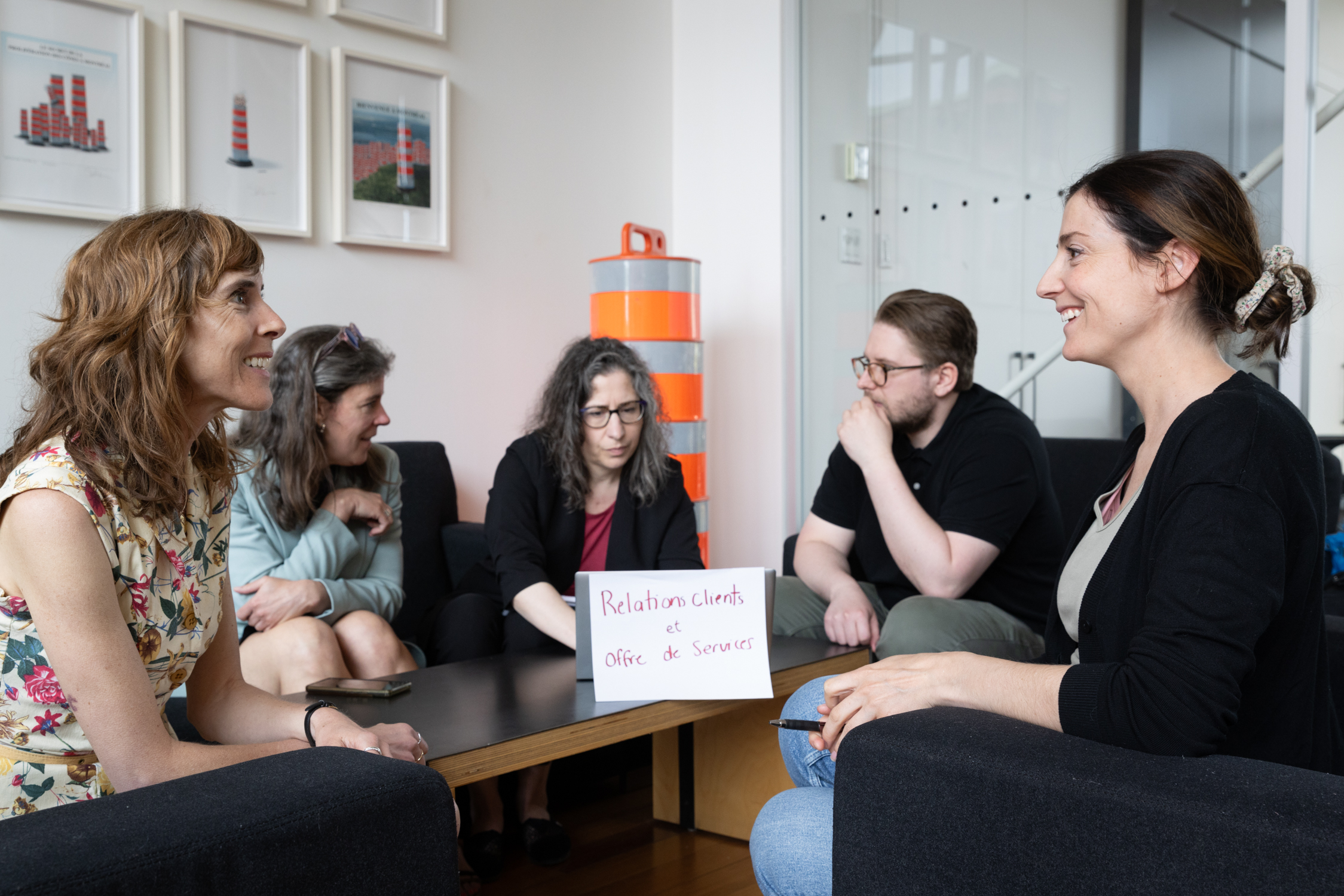 Cinq personnes sont assises dans un bureau et discutent. Au premier plan, deux femmes se font face, tandis que les trois autres sont assises autour d'une table sur laquelle est posé un panneau indiquant « Relations clients, Bureau de services ».
