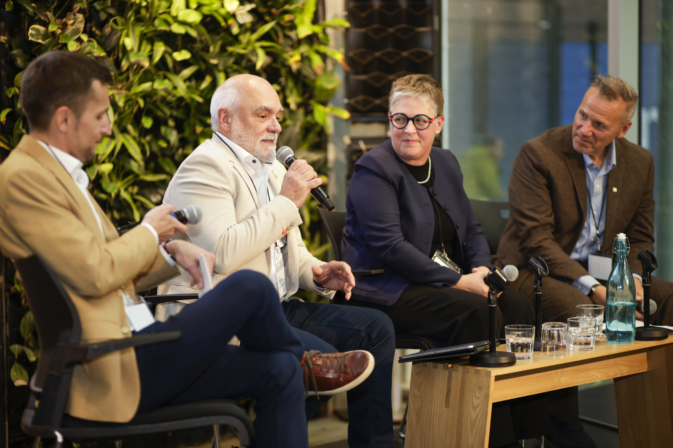 Quatre personnes en tenue professionnelle sont assises sur des chaises et s'expriment au micro lors d'une table ronde. Deux verres et une bouteille d'eau sont posés sur une table en bois devant elles. Des plantes vertes servent de toile de fond d'un côté.