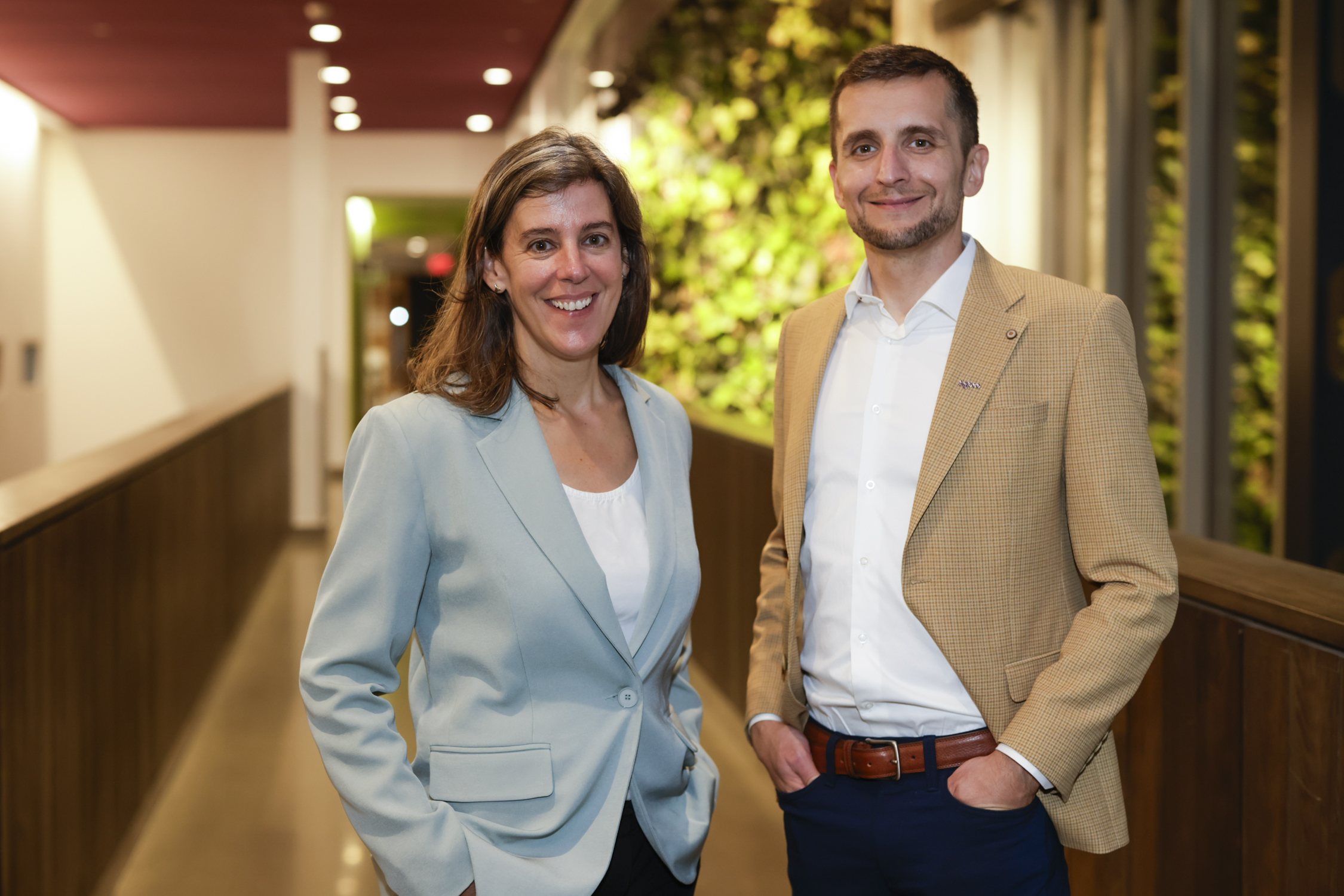 Deux personnes en tenue professionnelle se tiennent debout et sourient dans un couloir, avec en arrière-plan un mur végétal et un éclairage vif.
