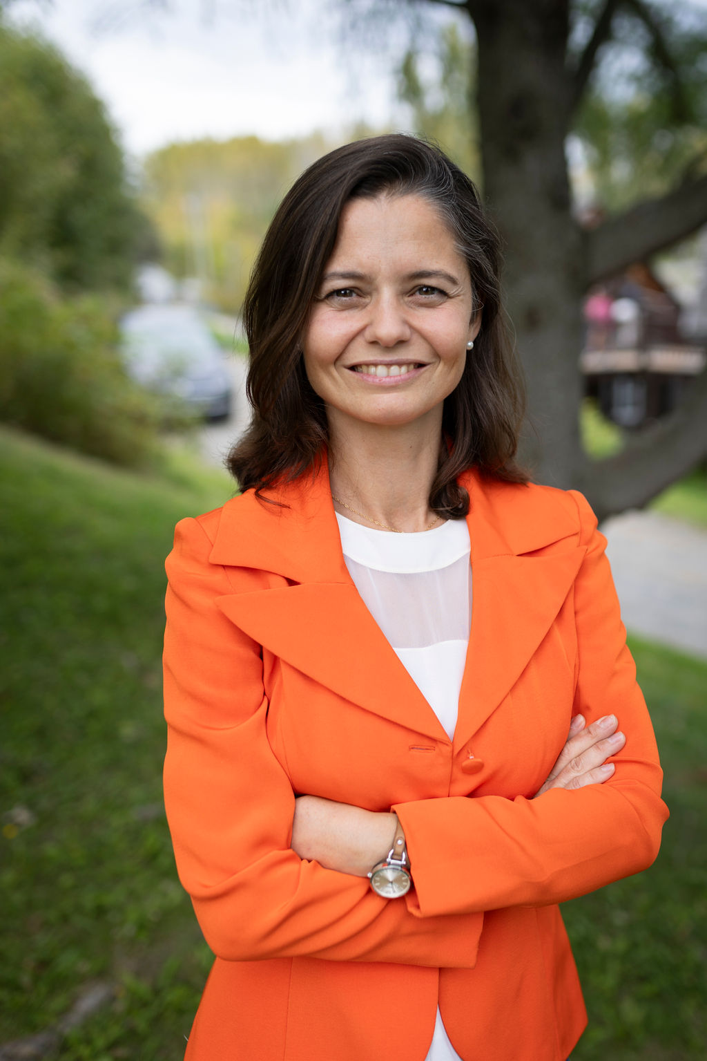 Une femme aux cheveux châtains, vêtue d'un blazer orange, se tient à l'extérieur, les bras croisés, et sourit à l'appareil photo. On aperçoit de l'herbe verte et des arbres en arrière-plan.