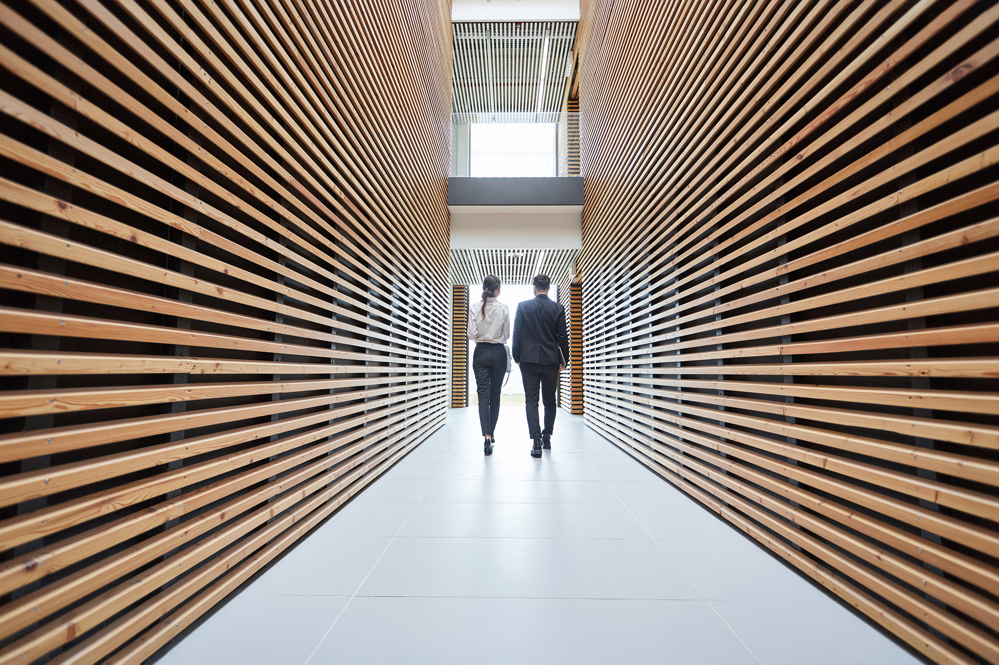 Deux personnes en tenue professionnelle avancent dans un couloir moderne bordé de lattes de bois horizontales, en direction d'une fenêtre lumineuse située au fond.