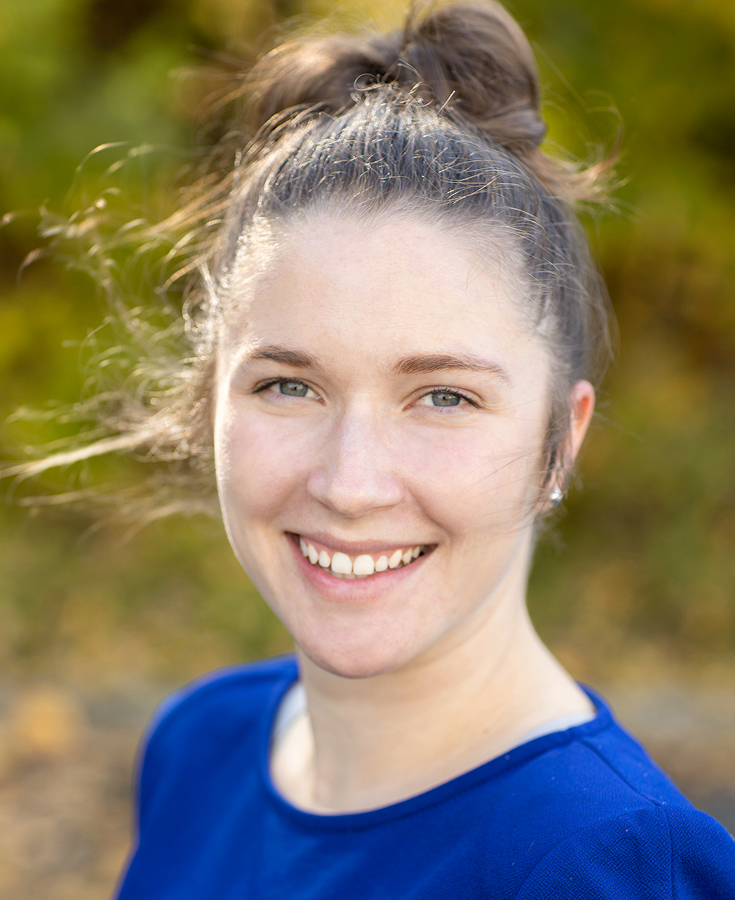 Une femme souriante, à la peau claire et aux cheveux bruns relevés en chignon décoiffé, vêtue d'un t-shirt bleu, se tient à l'extérieur, avec en arrière-plan un feuillage vert et jaune flou.