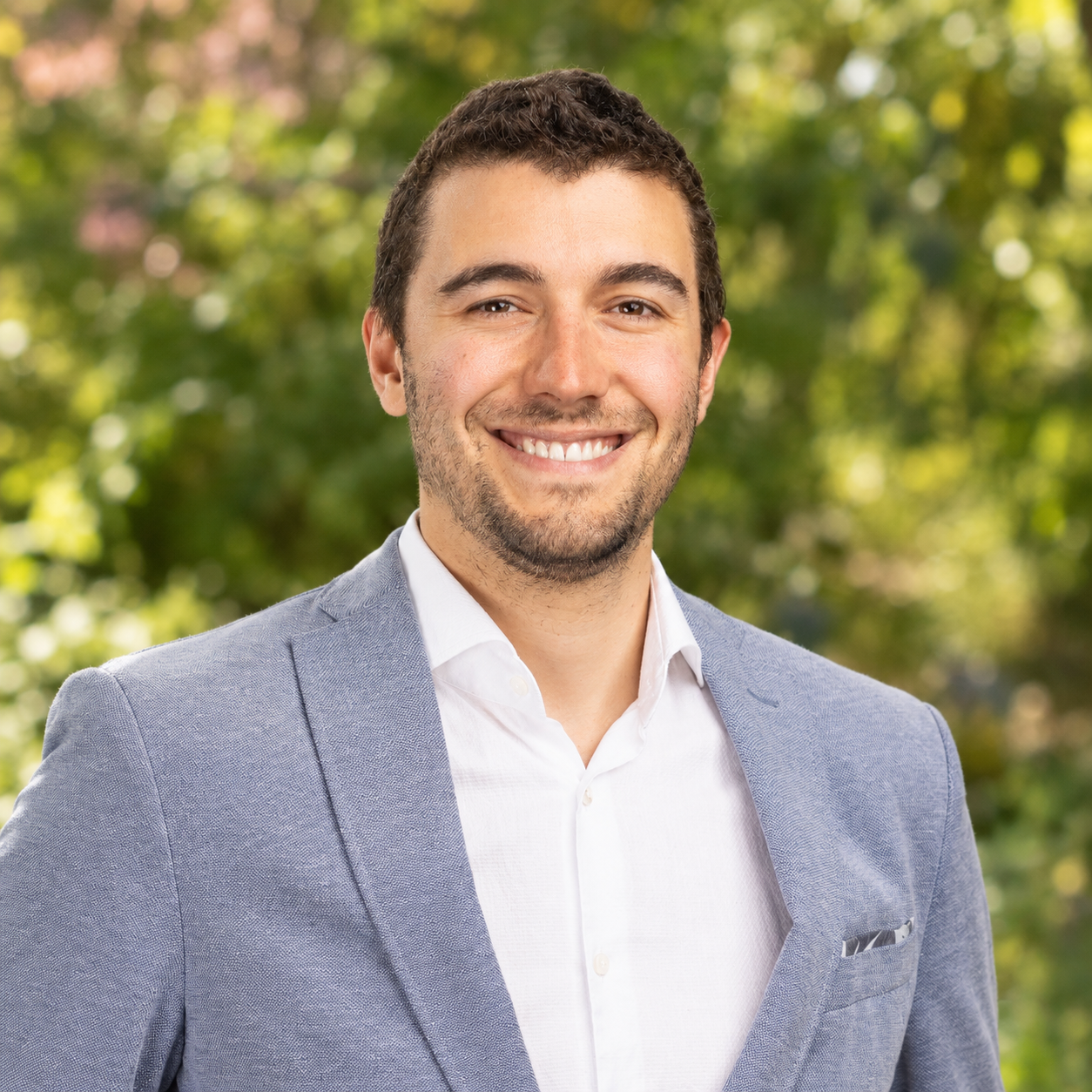 Un homme souriant, aux cheveux courts et foncés et à la barbe soignée, vêtu d'un blazer bleu clair et d'une chemise blanche, se tient debout à l'extérieur, avec des arbres verts flous en arrière-plan.