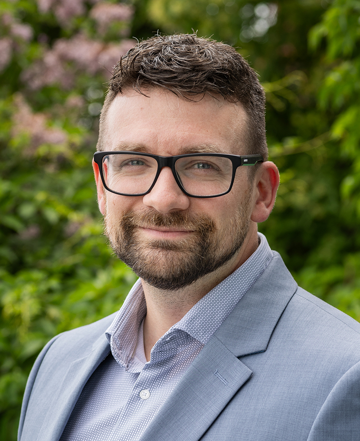 Un homme aux cheveux bruns courts, portant des lunettes et une barbe, est vêtu d'un blazer bleu clair et d'une chemise blanche ; il se tient debout à l'extérieur, avec en arrière-plan des arbres verts et des fleurs violettes.