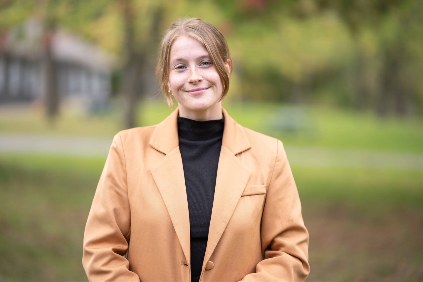 Une personne aux cheveux châtain clair relevés en chignon, vêtue d'un blazer beige et d'un haut noir, se tient à l'extérieur et sourit, avec une végétation floue en arrière-plan.