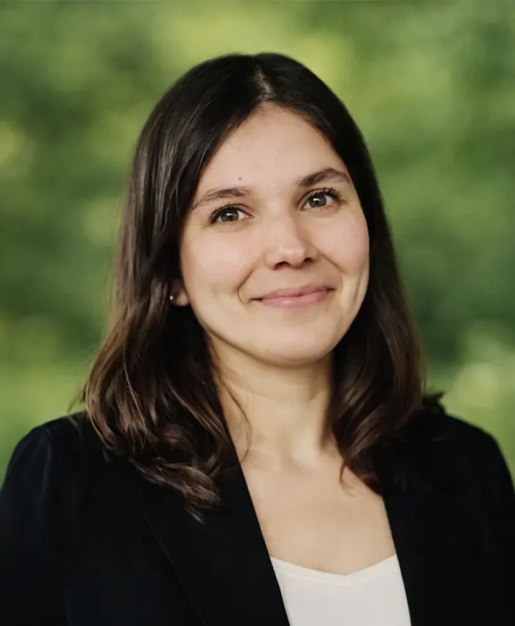 Une femme aux cheveux châtains mi-longs sourit doucement ; elle porte un blazer noir par-dessus un haut blanc. L'arrière-plan est constitué d'une végétation floue.
