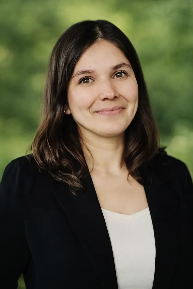 Une femme aux cheveux bruns mi-longs, vêtue d'un blazer foncé et d'un haut blanc, sourit doucement à la caméra sur fond de nature verdoyante et floue.