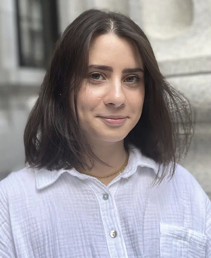 Une femme aux cheveux bruns mi-longs, vêtue d'une chemise blanche boutonnée et portant un collier en or, se tient à l'extérieur devant un bâtiment en pierre, souriant doucement à la caméra.
