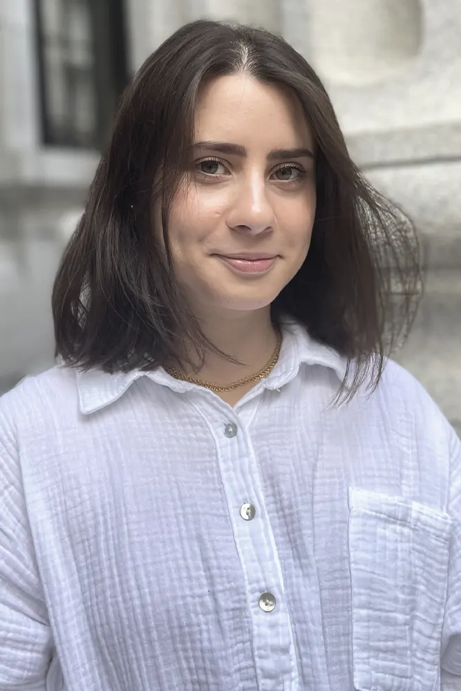 Une personne aux cheveux châtains mi-longs, vêtue d'une chemise blanche boutonnée et portant un collier en or, se tient à l'extérieur près d'un bâtiment en pierre, souriant doucement à la caméra.