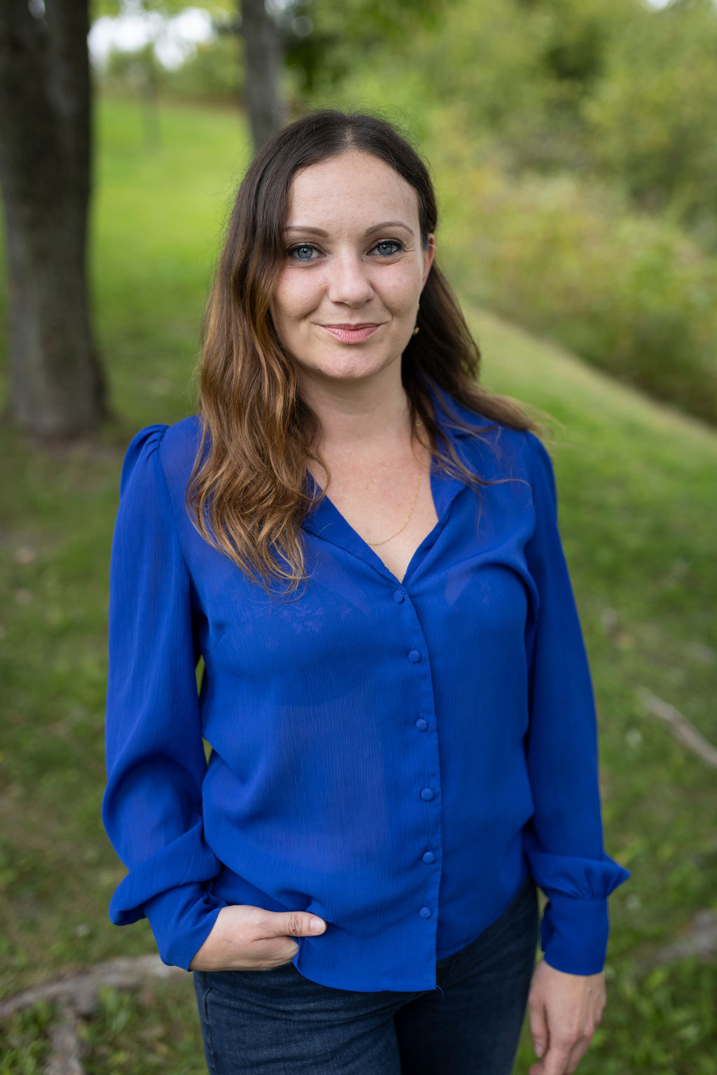 Une femme aux cheveux bruns ondulés, vêtue d'une chemise bleue boutonnée et d'un jean, se tient debout à l'extérieur sur une pelouse verte, avec des arbres et des buissons en arrière-plan. Elle sourit et a une main dans sa poche.