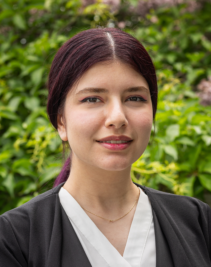 Une femme aux cheveux noirs et raides, vêtue d'un blazer noir par-dessus un haut blanc, se tient à l'extérieur devant un feuillage verdoyant, souriant légèrement à la caméra.